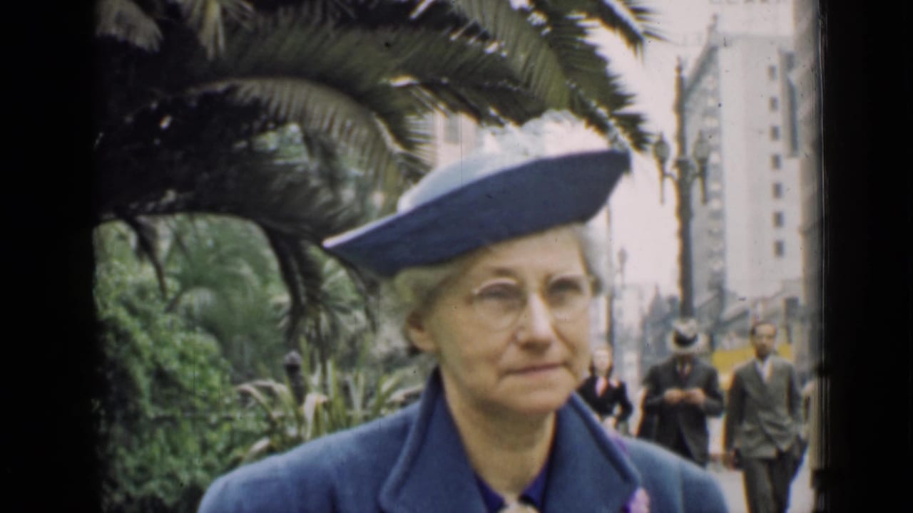 Older woman in a blue hat and coat walking near palms and city buildings.