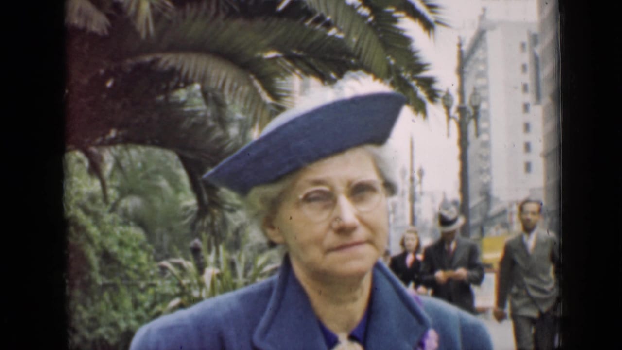 Older woman in blue formal attire facing the camera near palm trees.