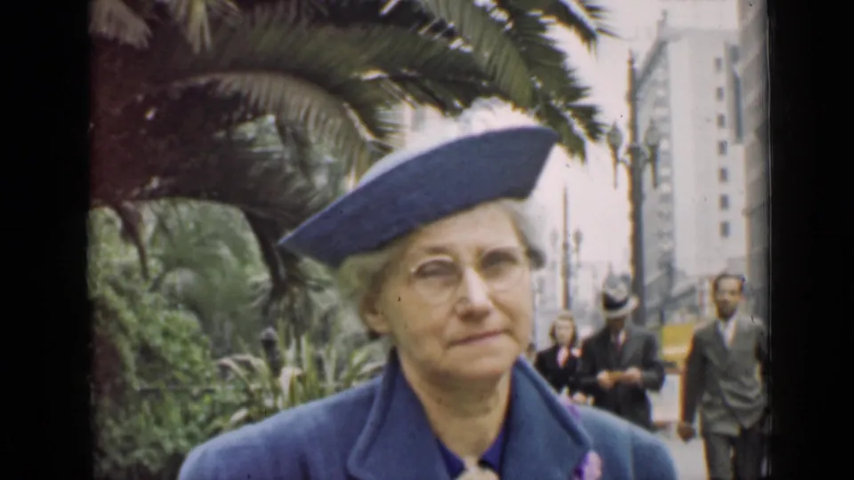 The woman in a blue hat and coat faces the camera, with pedestrians and tall buildings in the background.