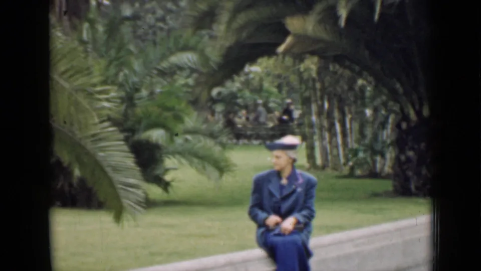 The woman sits on a low border in the urban park, wearing her blue hat and coat amid lawn and palm plantings.