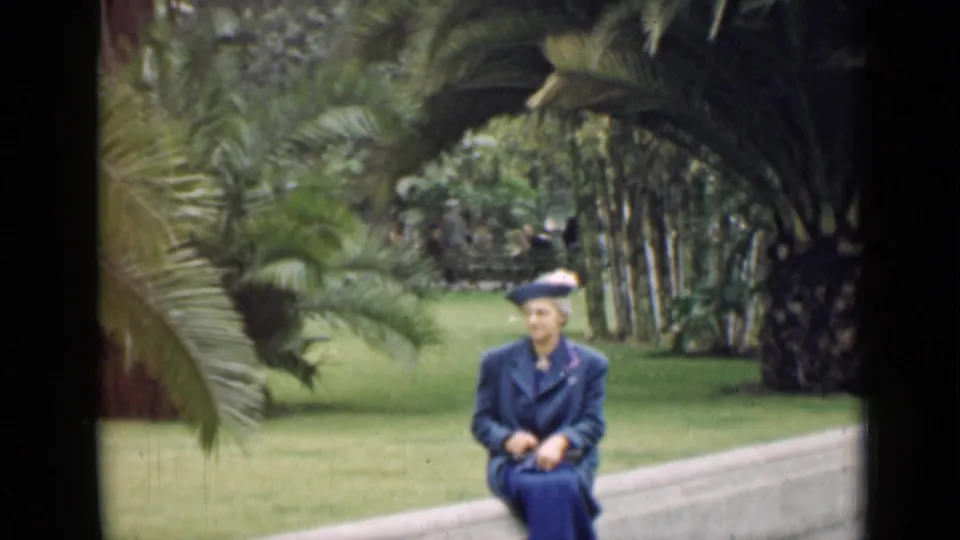 1939 Arizona 8mm frame showing a woman in a blue coat and hat seated on a concrete ledge amid palms and lawn.