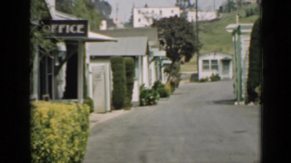 1939 Arizona 8mm view of a quiet paved drive lined by white buildings, an office sign, hedges, utility poles, and a green hillside.