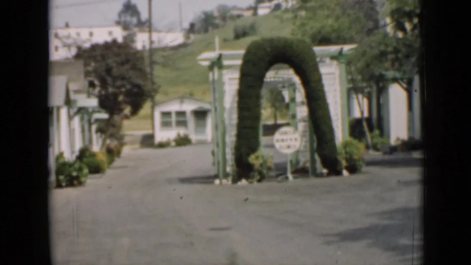 1939 Arizona home-movie frame showing a paved entrance with an arched topiary, small sign, white buildings, hedges, and hillside beyond.