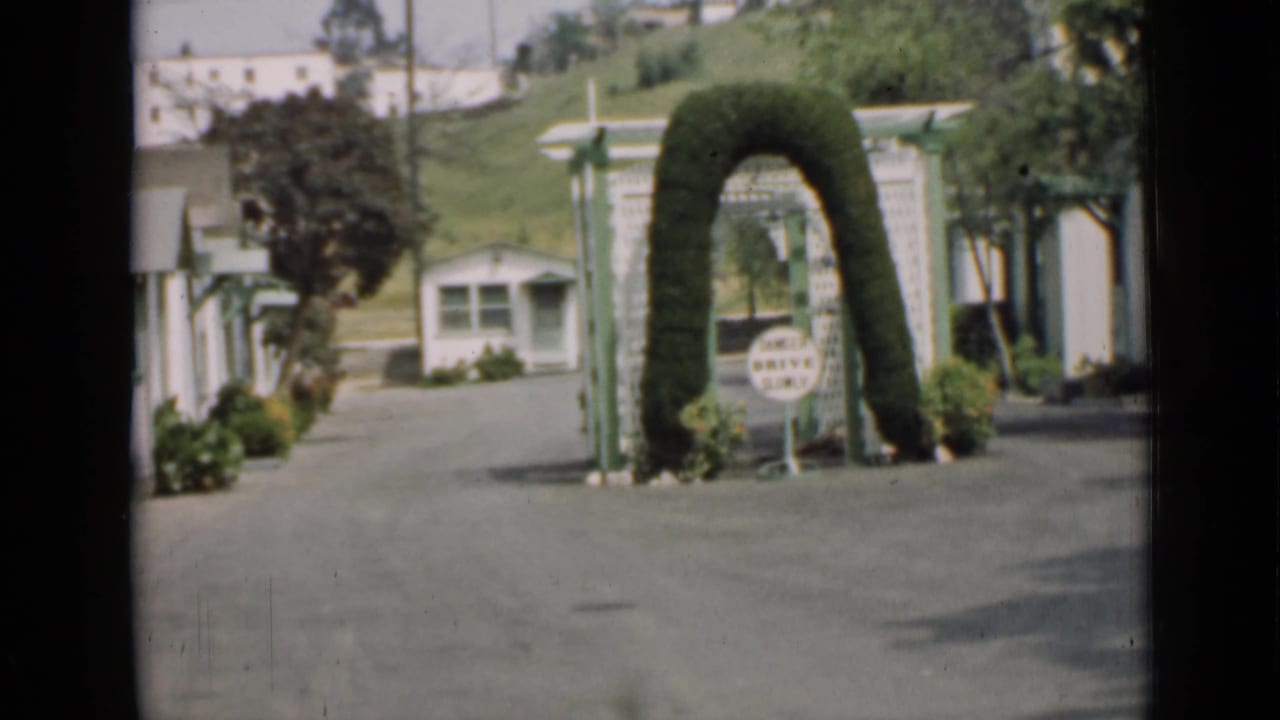 Paved lane between small buildings with a trimmed arched hedge and a small sign.