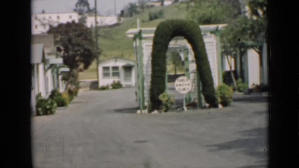 1939 Arizona home-movie frame showing a quiet lane, small white buildings, landscaped shrubs, a green arched hedge, and a small sign.