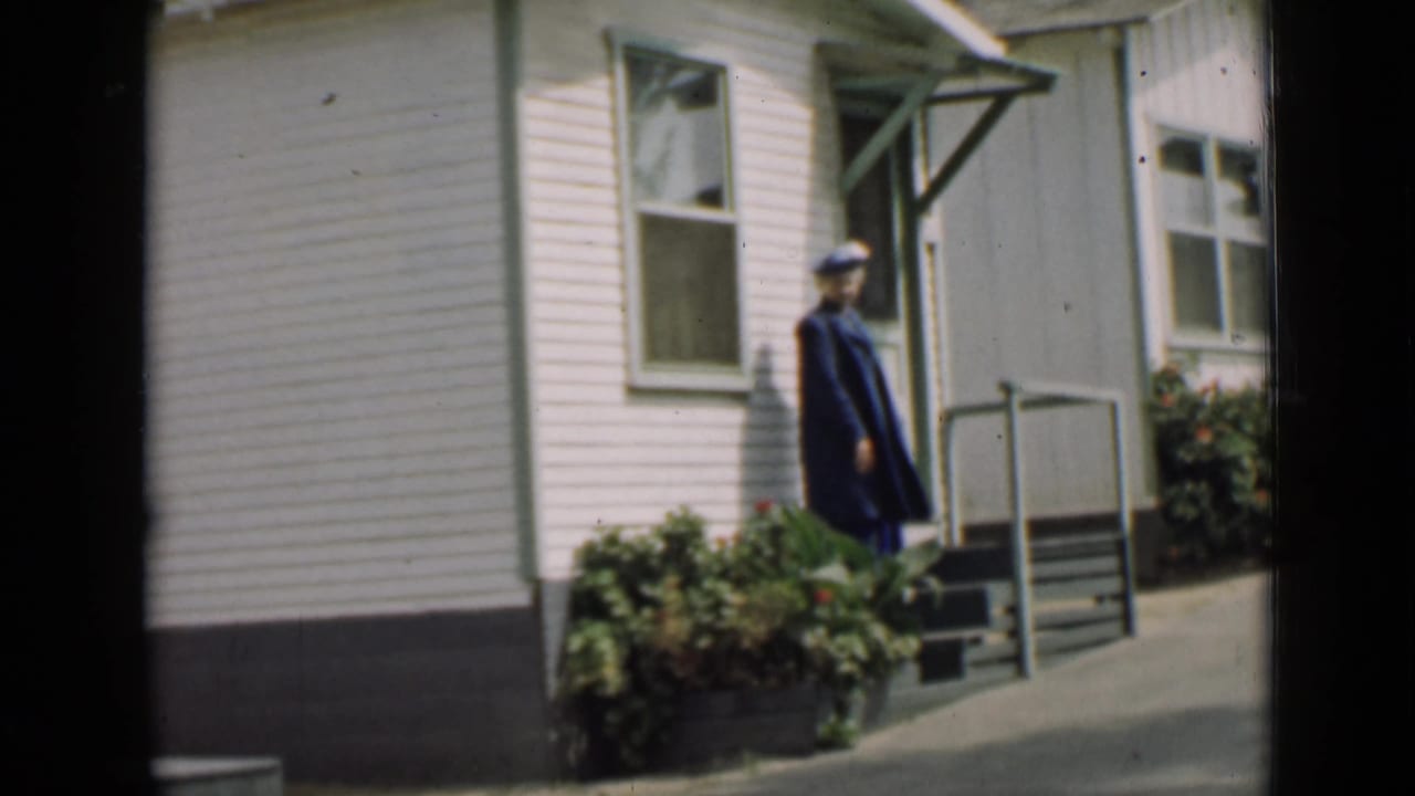 Uniformed adult in a dark coat and cap stands beside a white-sided building porch.