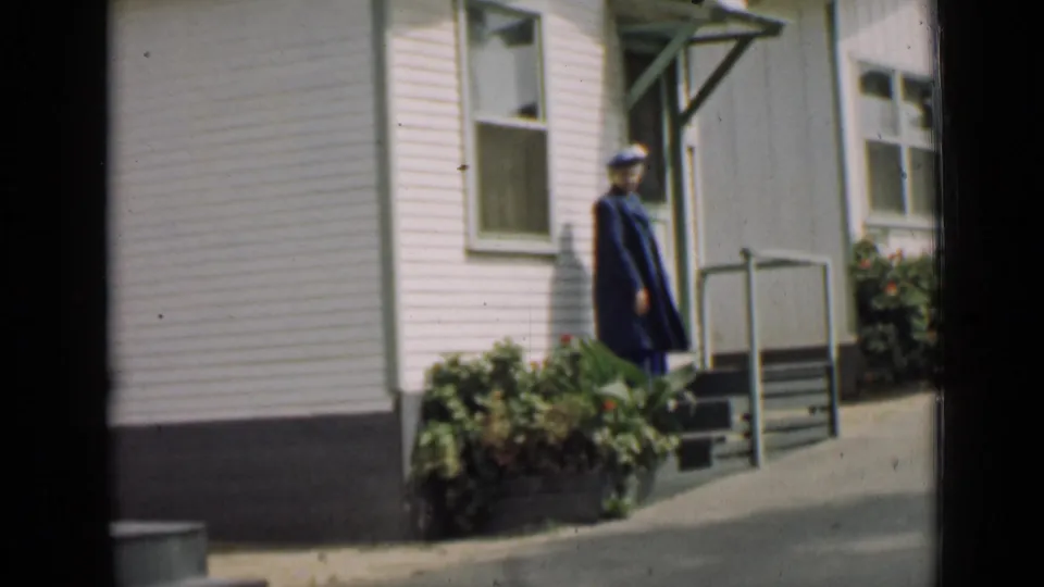 1939 Arizona frame showing a uniformed adult near a small porch, with white siding, railings, window, and shrubs around the entrance.