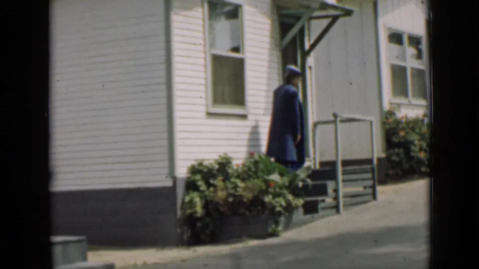 1939 Arizona frame showing a uniformed adult turning near the steps of a white-sided building, with shrubs and railings in the foreground.