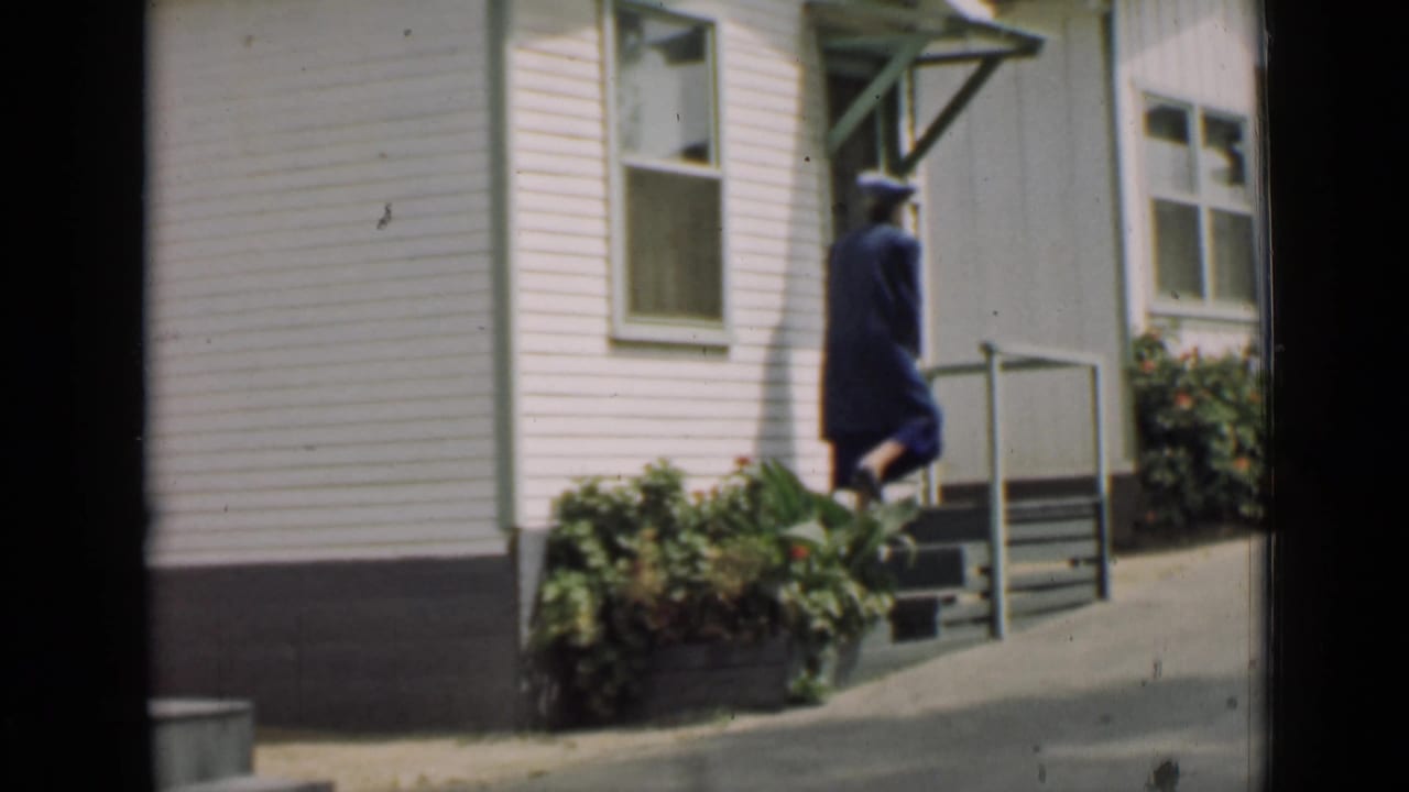 Woman in a blue dress walks toward steps beside a white clapboard house.