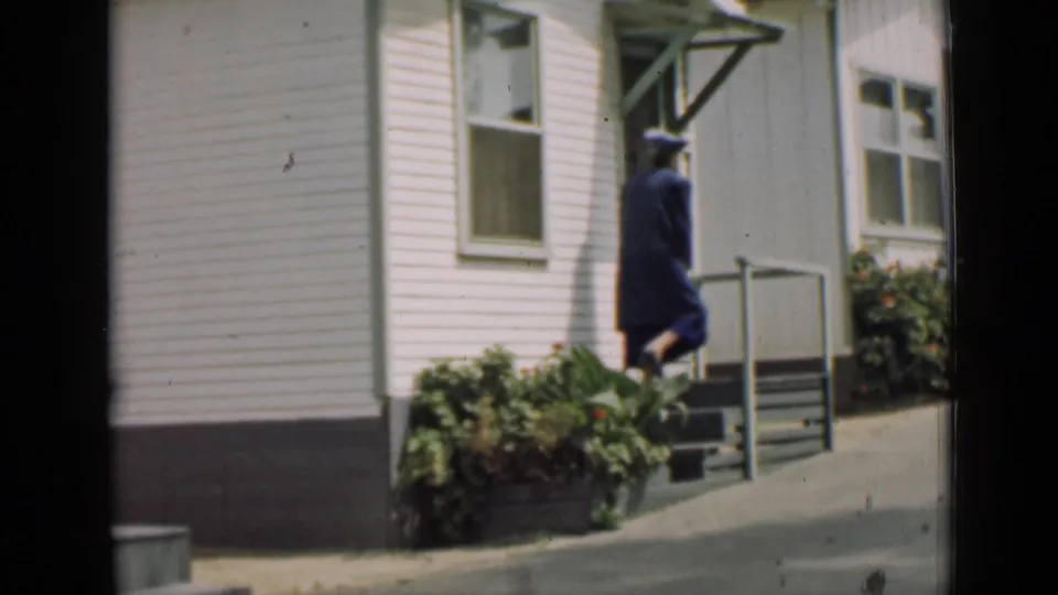 In Arizona 1939 footage, a woman in a blue dress and hat approaches porch steps beside a white clapboard house.