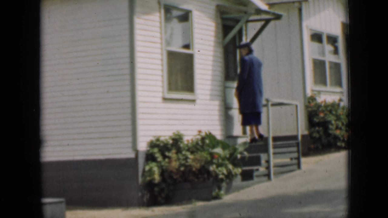 Woman stands near the doorway at the top of the steps of a white house.