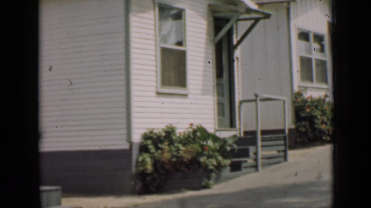 White-sided house exterior with green trim, porch steps, railings, driveway, and shrubs.