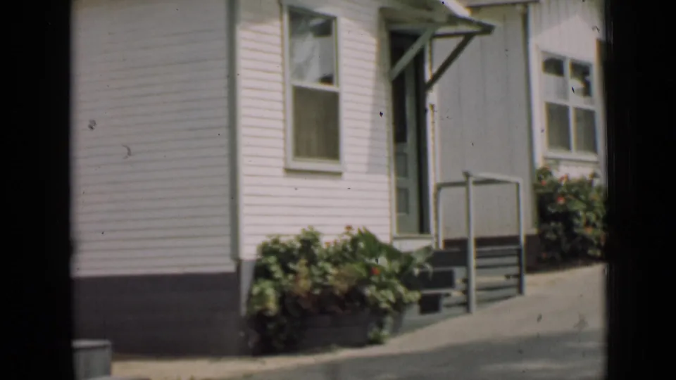 1939 Arizona 8mm frame of a modest white-sided house with green trim, porch steps, railings, driveway, and planted shrubs.