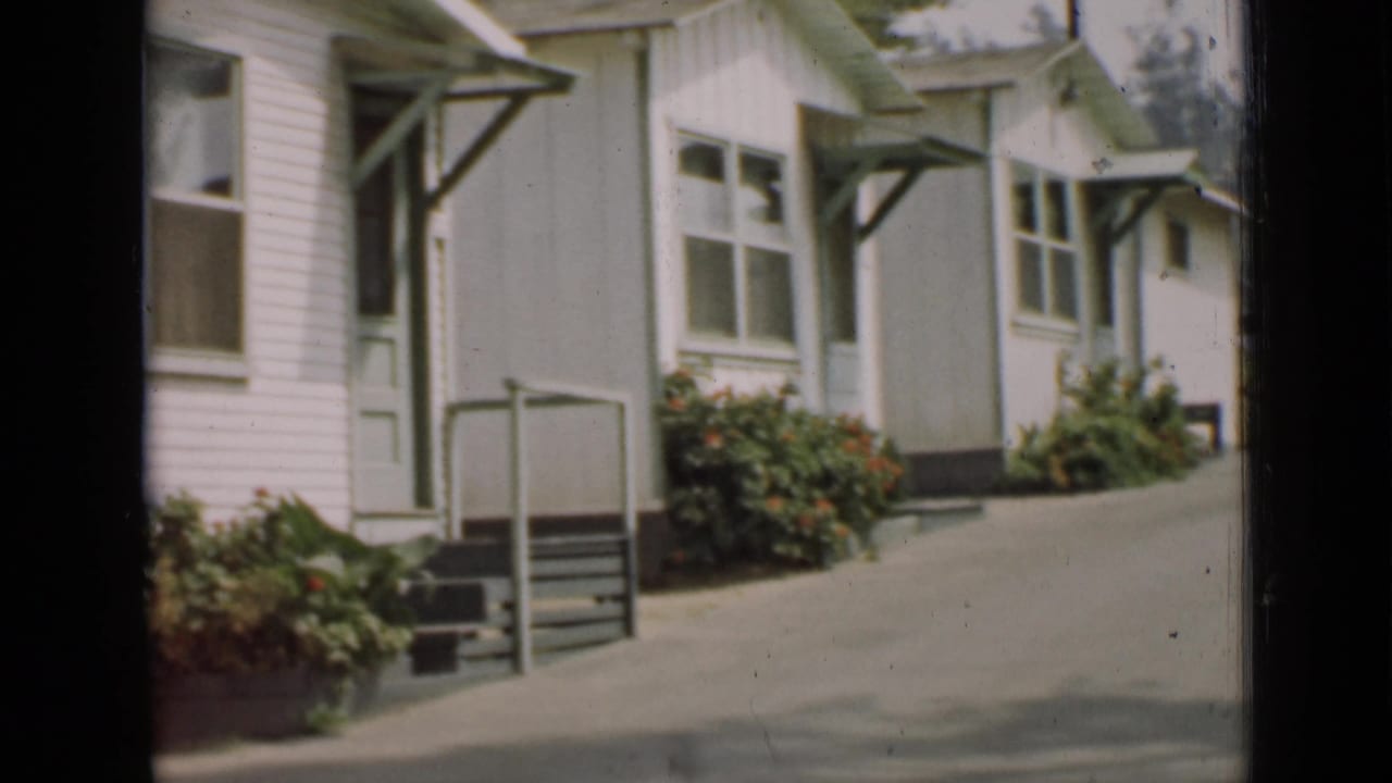 Row of similar white houses with green trim, small porches, paved drive, and flowerbeds.