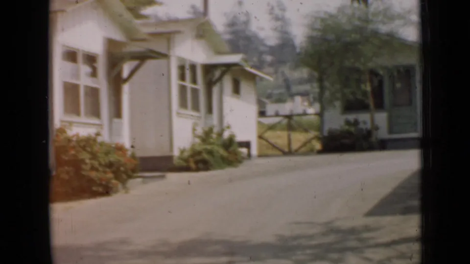 1939 Arizona home-movie frame with a house corner, shrubs, paved drive, gate, trees, and a small outbuilding or shed beyond.