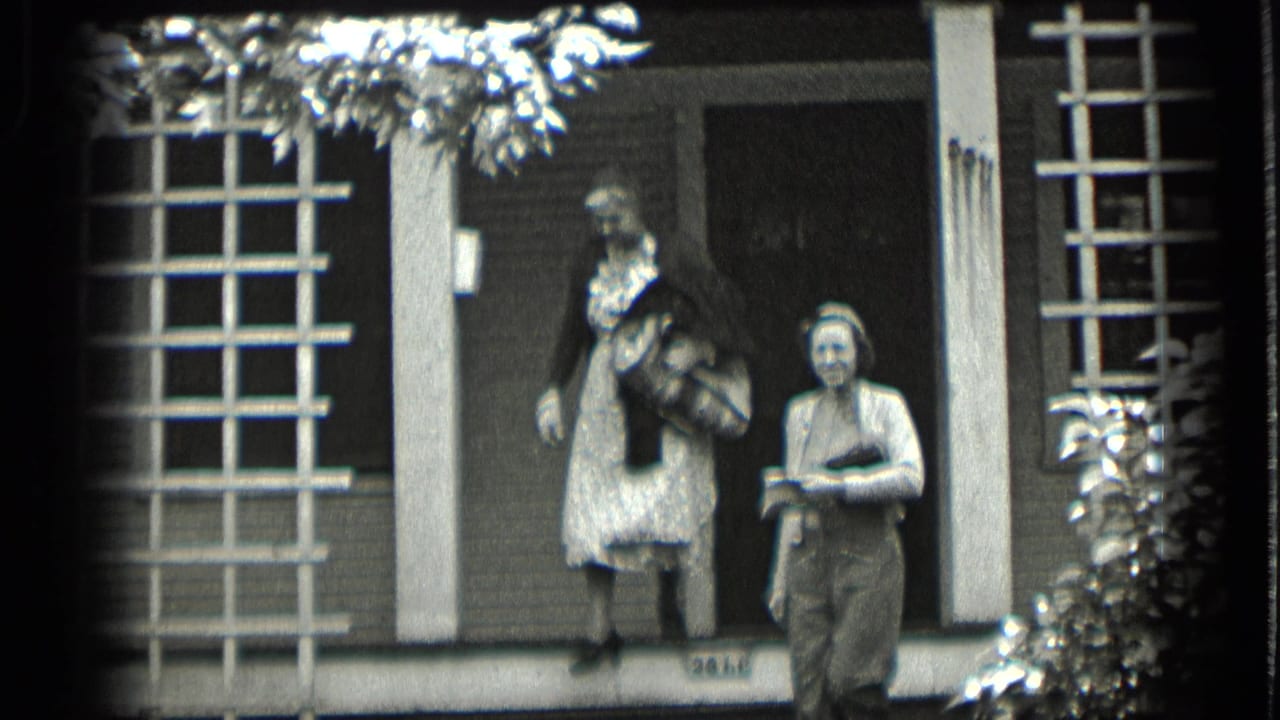 In 1939 Clear Lake home-movie footage, a woman in a dark sweater and patterned dress stands on porch steps while another woman waits nearby.