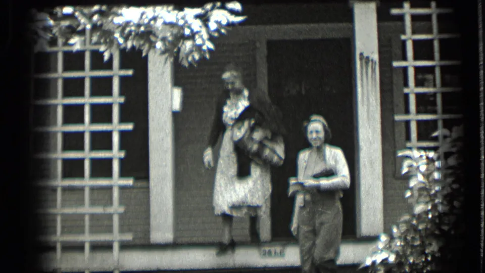 In 1939 Clear Lake home-movie footage, a woman in a dark sweater and patterned dress stands on porch steps while another woman waits nearby.