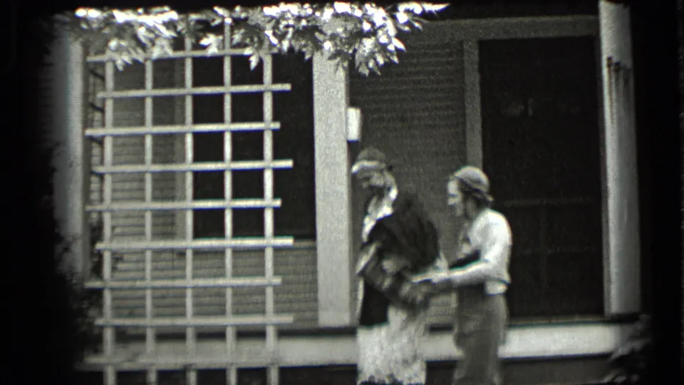Two women appear to exchange or handle items on a Clear Lake porch, with a gridded window and house siding behind them.