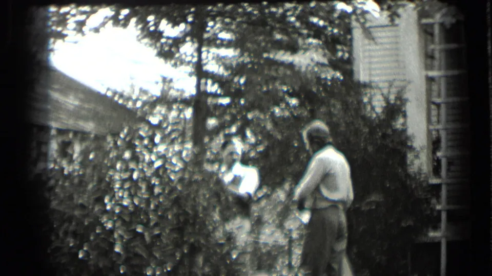 Two women continue through a shaded yard beside the Clear Lake house, partly obscured by shrubs and overhanging branches.