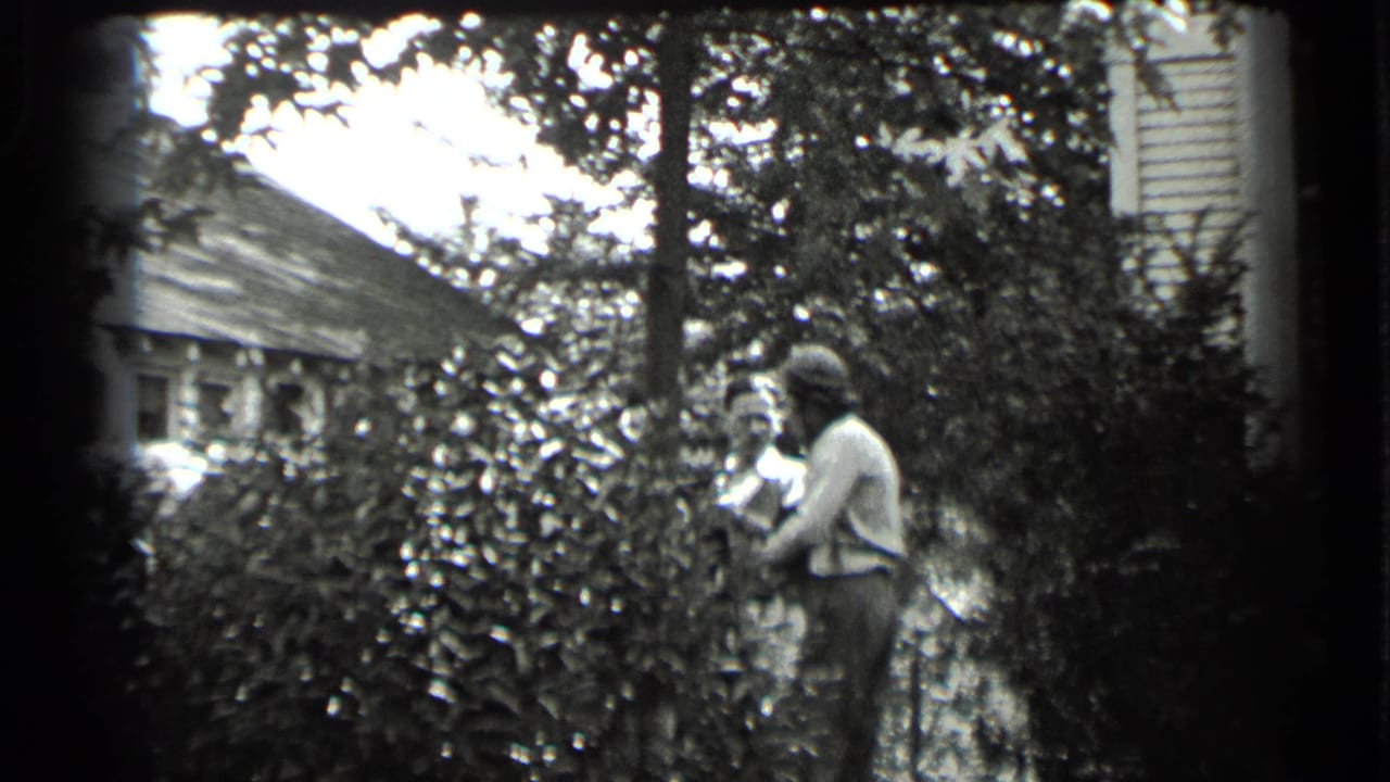 Two adults partly obscured by shrubs near a residential yard and neighboring houses.