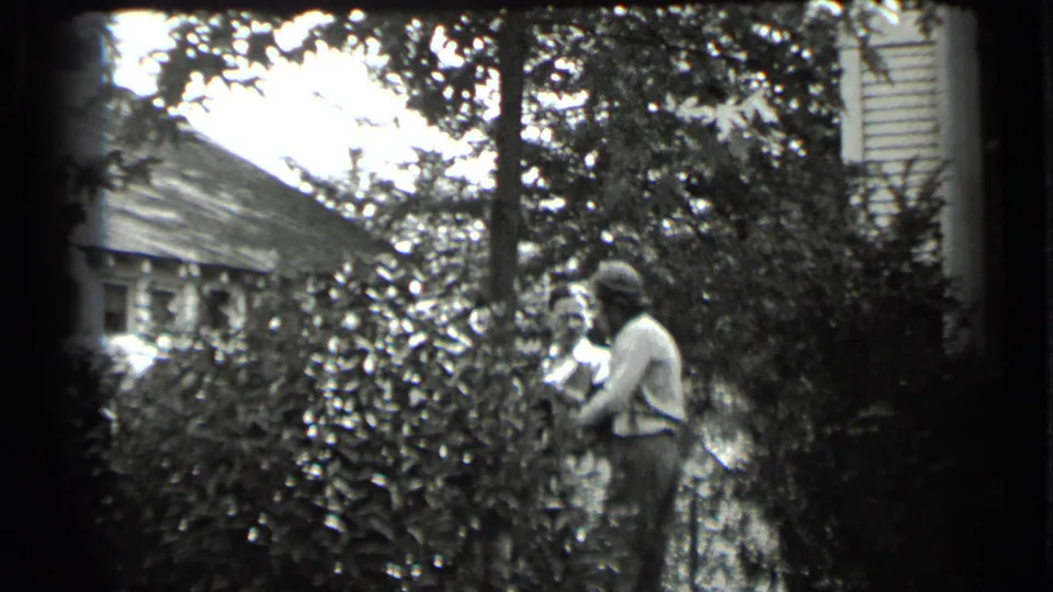 In Clear Lake, California home-movie footage from 1939, two adults stand or move near dense shrubs outside a residential house.