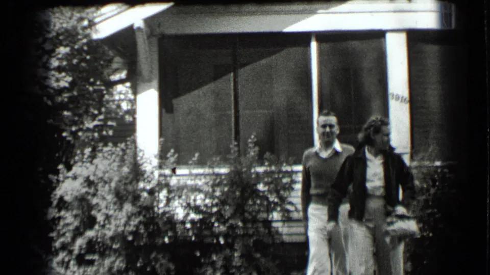 A man and woman walk side by side in front of the Clear Lake house, with the porch screen and garden plantings behind them.