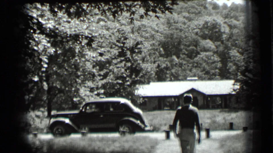 A man moves toward a rounded-body vintage car near trees and a low building in the supplied 1939 footage.