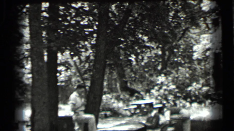 1939 Clear Lake home-movie frame showing adults relaxing near picnic benches in a shaded wooded area.