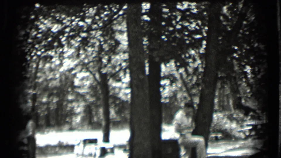 1939 Clear Lake frame with a seated adult near a picnic table, framed by trunks and dappled light.
