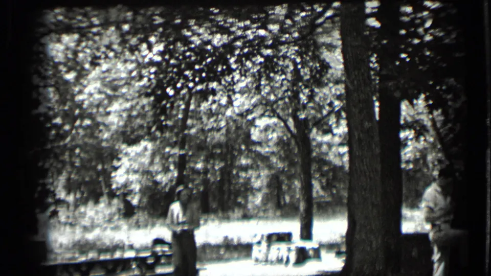 1939 Clear Lake home-movie frame showing adults standing in a shaded picnic grove with benches nearby.