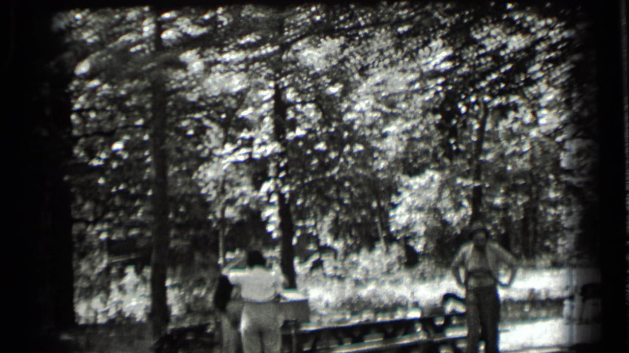 Two adults near a wooden picnic table in a shaded forest clearing.
