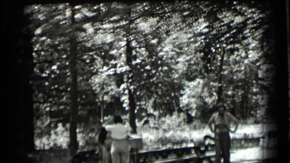 1939 Clear Lake home-movie frame showing two adults beside a picnic table under tall trees in dappled light.