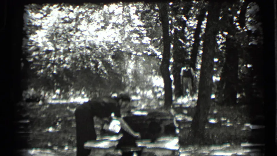 Black-and-white 8mm frame from Clear Lake in 1939 showing an adult leaning over a picnic table in the forest.