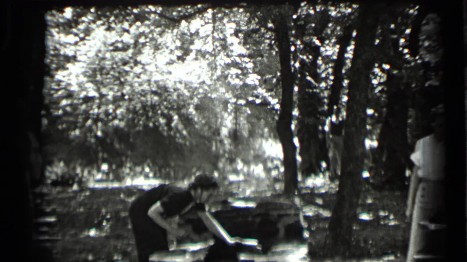 1939 Clear Lake home-movie frame showing picnic preparation under trees, with one adult bending at a table and another partly visible nearby.