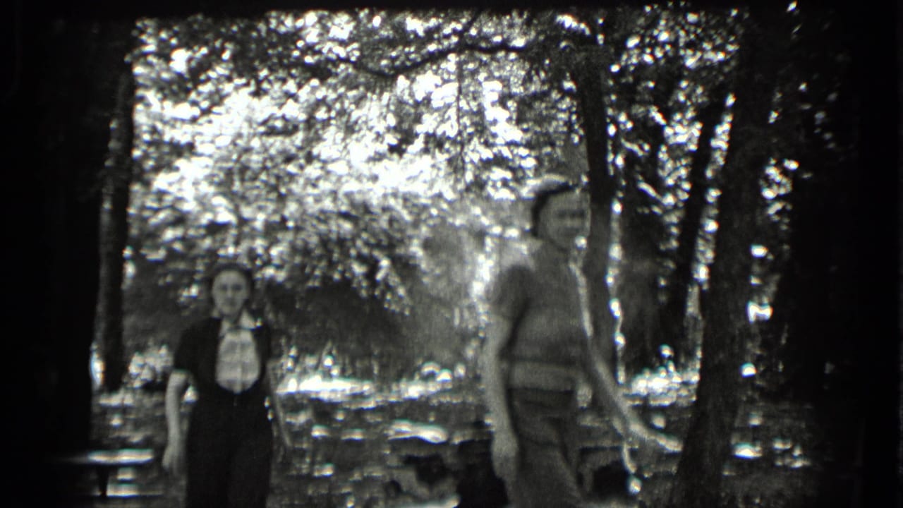 1939 Clear Lake frame with a young woman facing the camera and a young man walking past in a wooded lakeside picnic area.