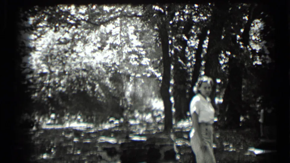 1939 Clear Lake frame with a woman in light clothing turning toward the camera beside trees and bright foliage.