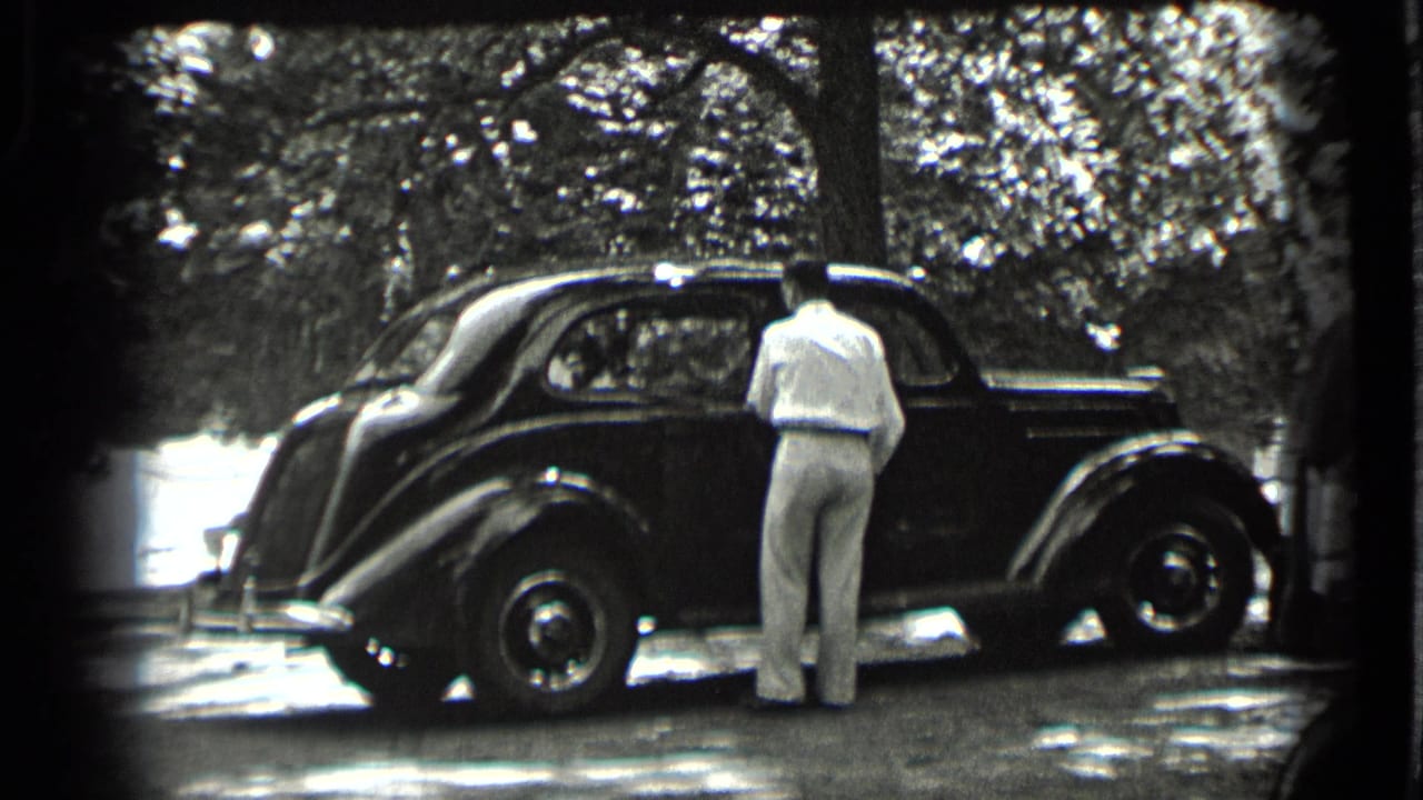 A man in light clothing stands next to a dark rounded sedan in a wooded Clear Lake setting, seen in black-and-white 8mm footage.