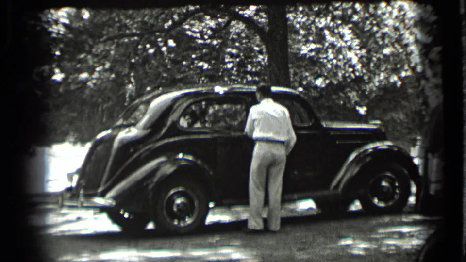 A man in light clothing stands next to a dark rounded sedan in a wooded Clear Lake setting, seen in black-and-white 8mm footage.