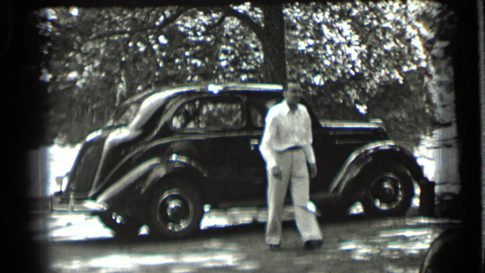The man faces the camera near the side of a dark 1930s-style sedan, with trees and bright patches of sunlight behind him.