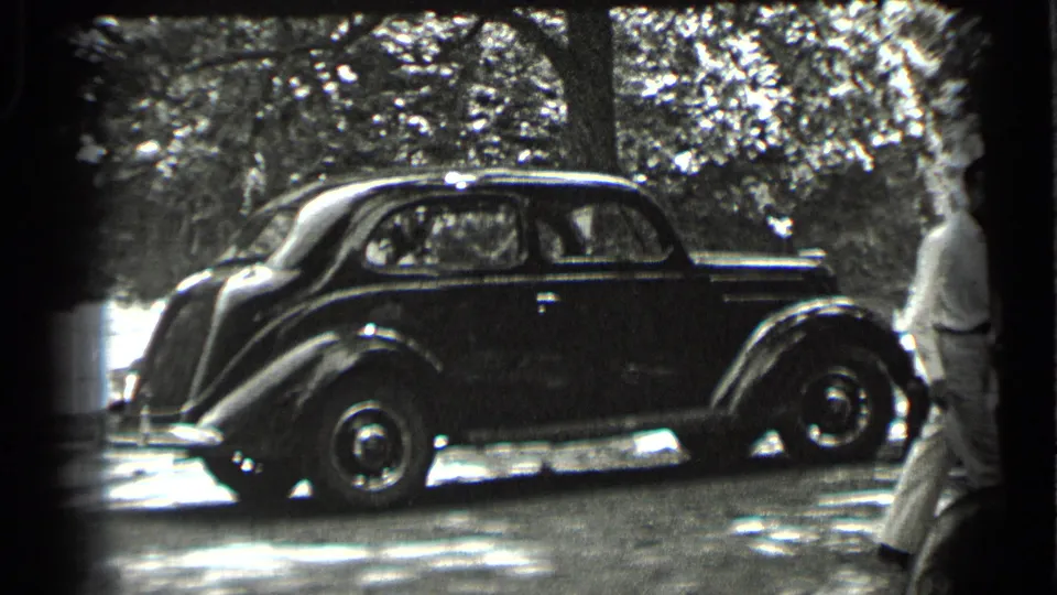 A dark rounded sedan remains parked beneath trees while the man in light clothing moves out of the right edge of the frame.