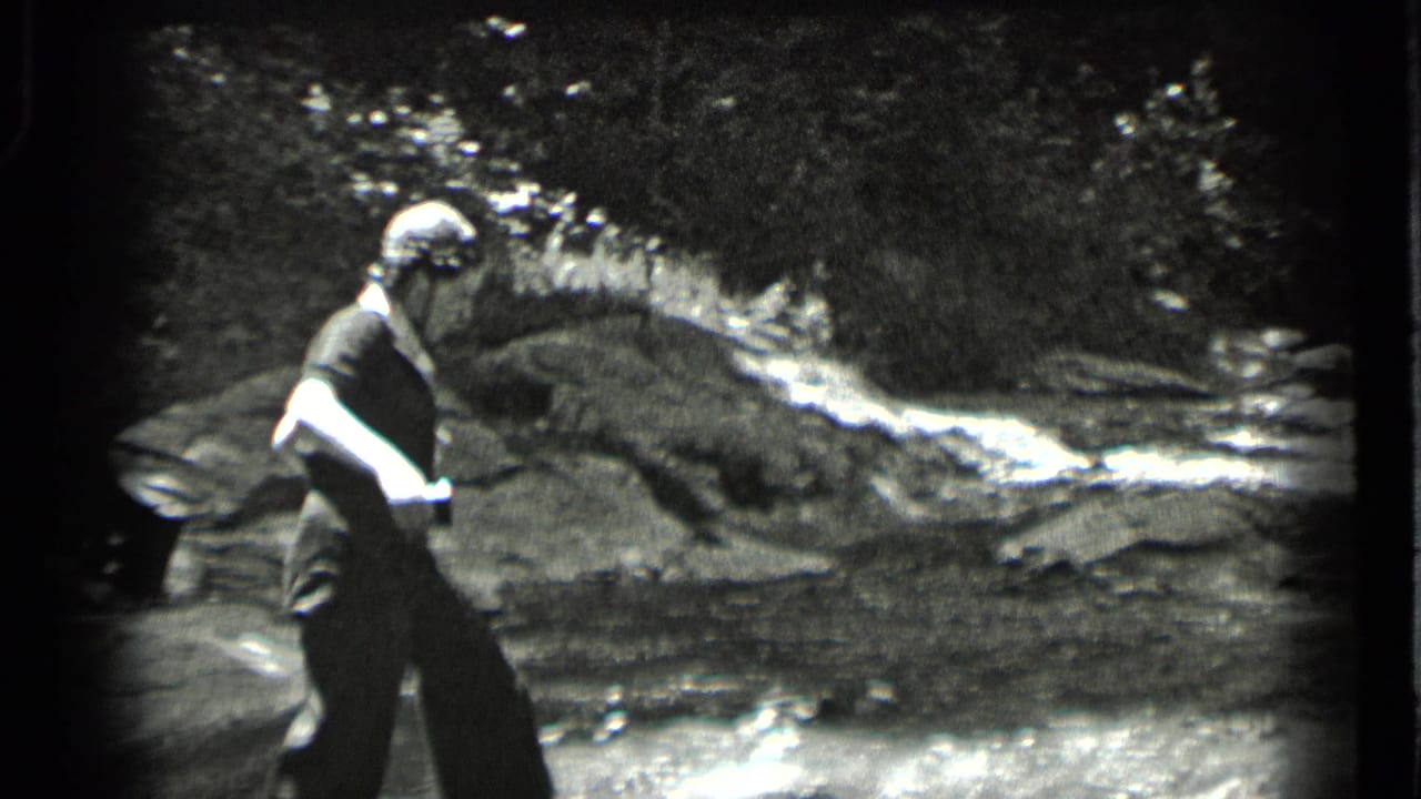 Woman in a light hat walks beside a rocky stream in a wooded area.