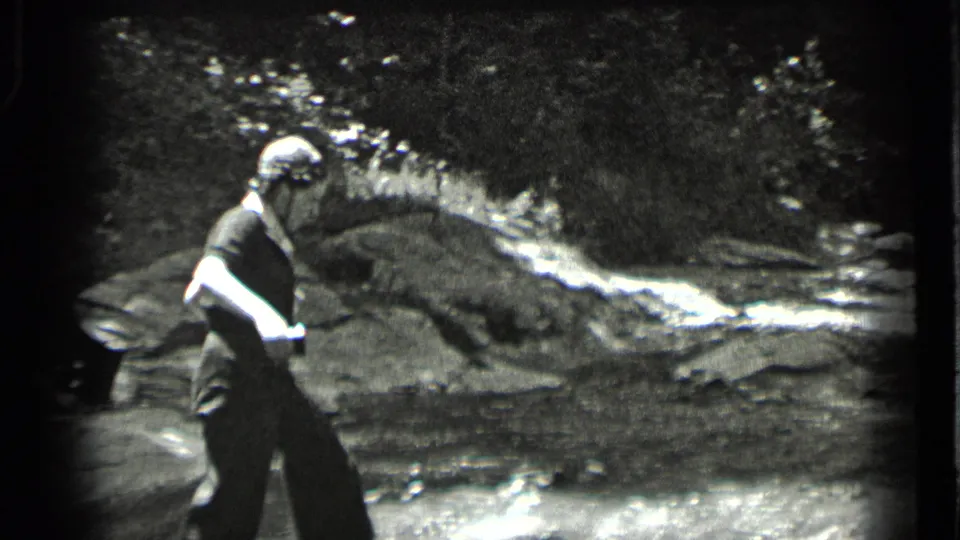 1939 Clear Lake home-movie frame showing a woman in a light hat moving along a rocky streambank in a wooded setting.