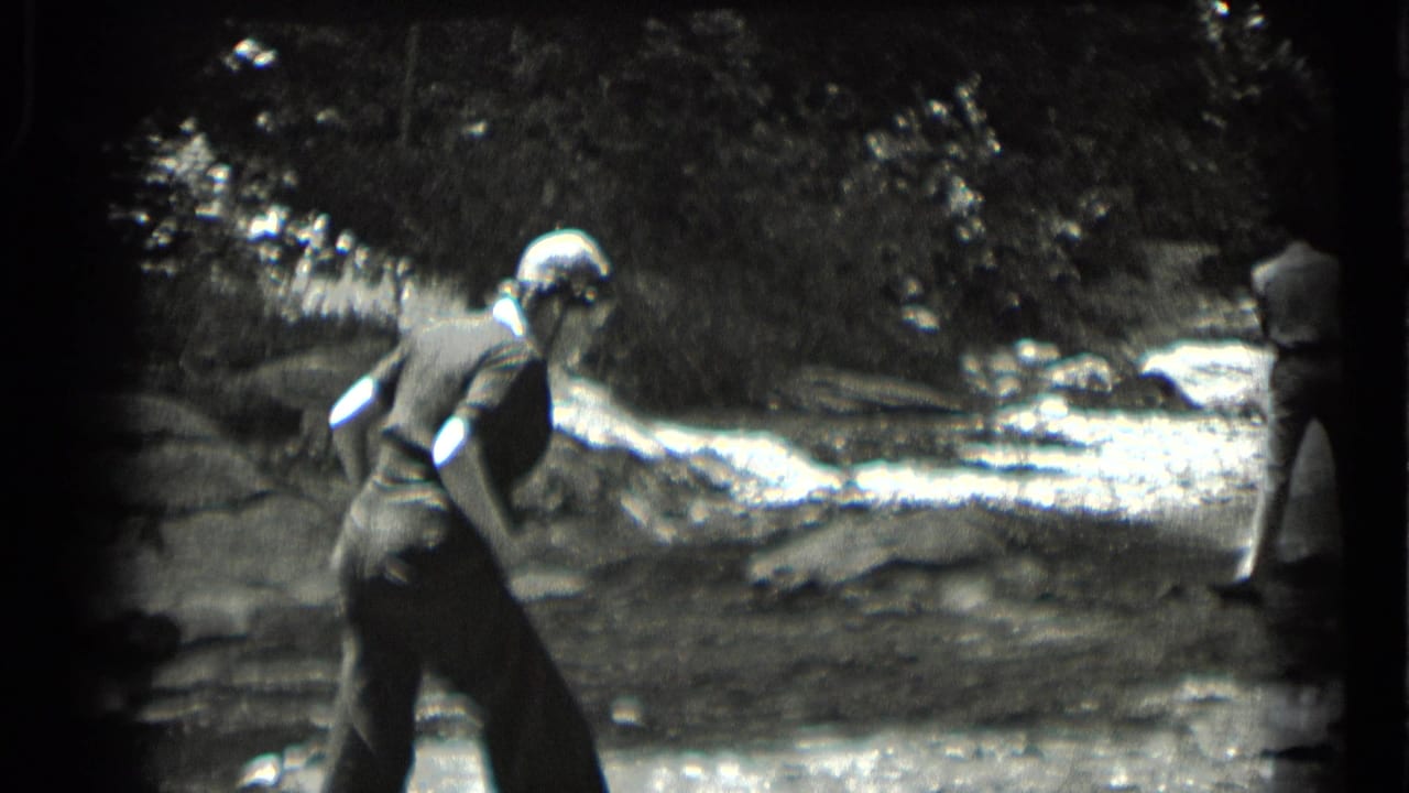 Woman bends slightly near a rocky stream while another adult stands at the right edge.