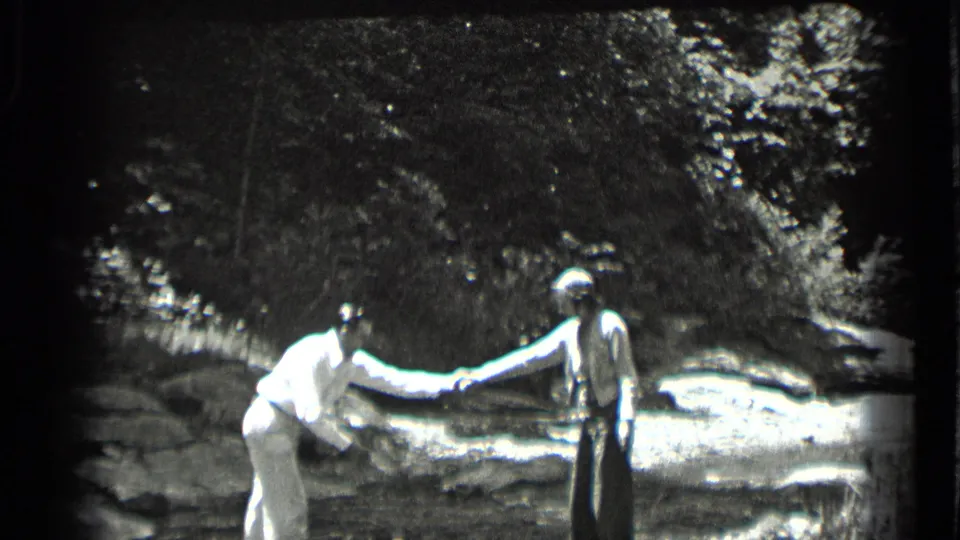 A woman in light clothing reaches toward a man standing on rocks beside the shallow Clear Lake stream.