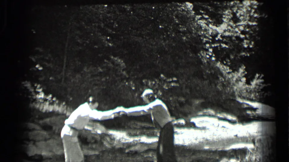 In a 1939 Clear Lake home-movie frame, two adults hold hands as one leans across the rocky stream.