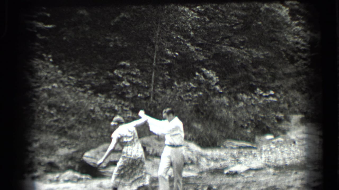 Man in light clothes helps a woman step across a shallow creek near rocks and trees.