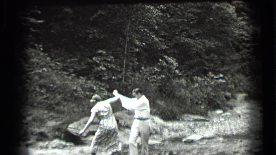At Clear Lake, California in 1939, a man steadies a woman in a patterned dress as she steps across a shallow wooded creek.