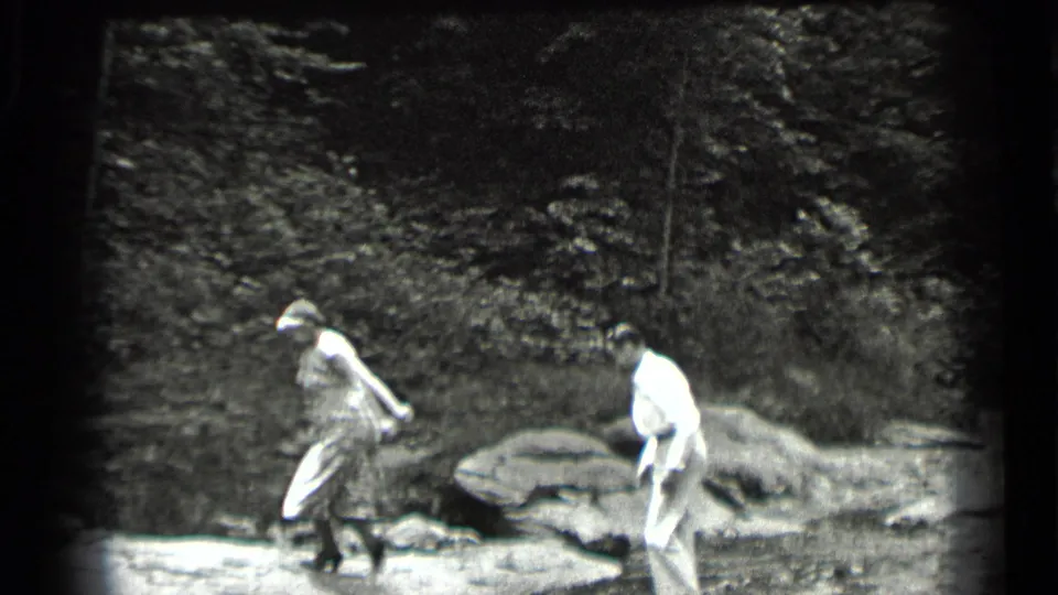 A woman in a dress and hat steps across the creek at Clear Lake while a man in light trousers follows behind her.
