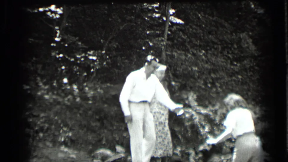 A man in a white shirt reaches toward a woman at the creek edge while another woman stands partly behind him in the wooded setting.