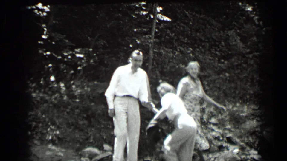 1939 Clear Lake home-movie frame showing a man and two women outdoors, one woman bending as the group stands near trees and brush.